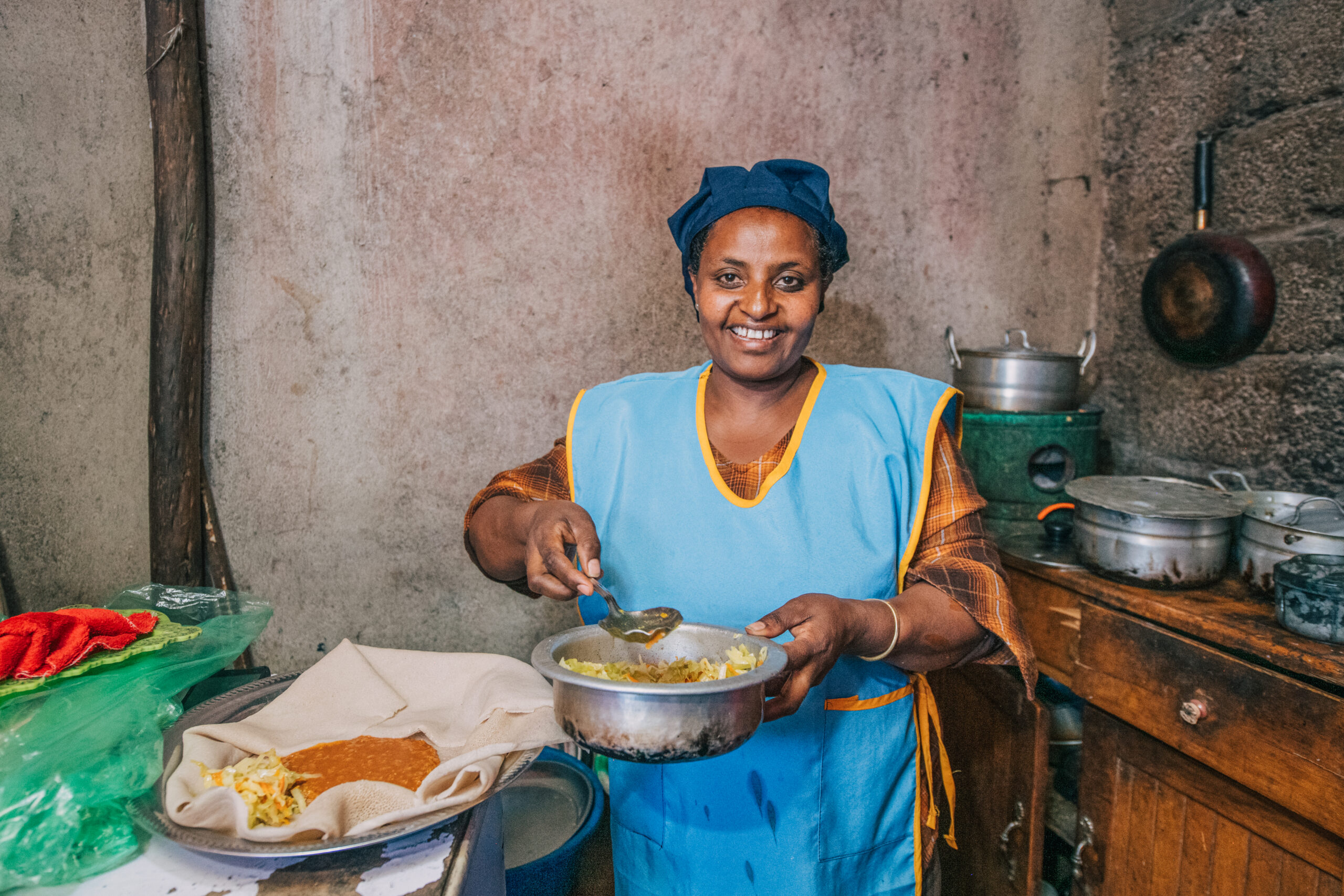 A smiling Ethiopian woman wearing an apron and hair cover proudly preparing and serving traditional food for her independent, widow-owned culinary business.