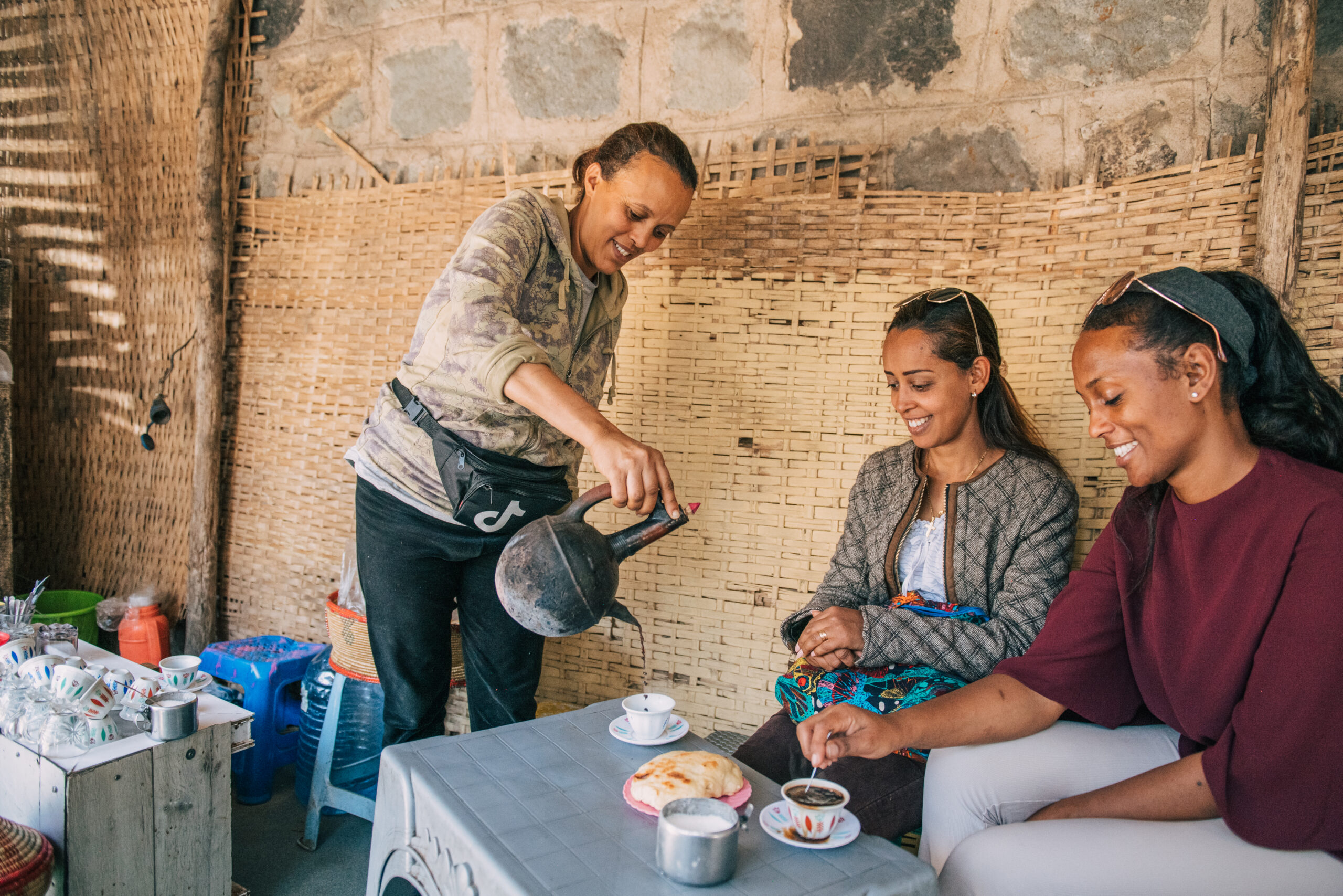 Yodit pouring traditional Ethiopian coffee from a jebena for two seated guests at her coffee shop, surrounded by traditional cups and warm afternoon light